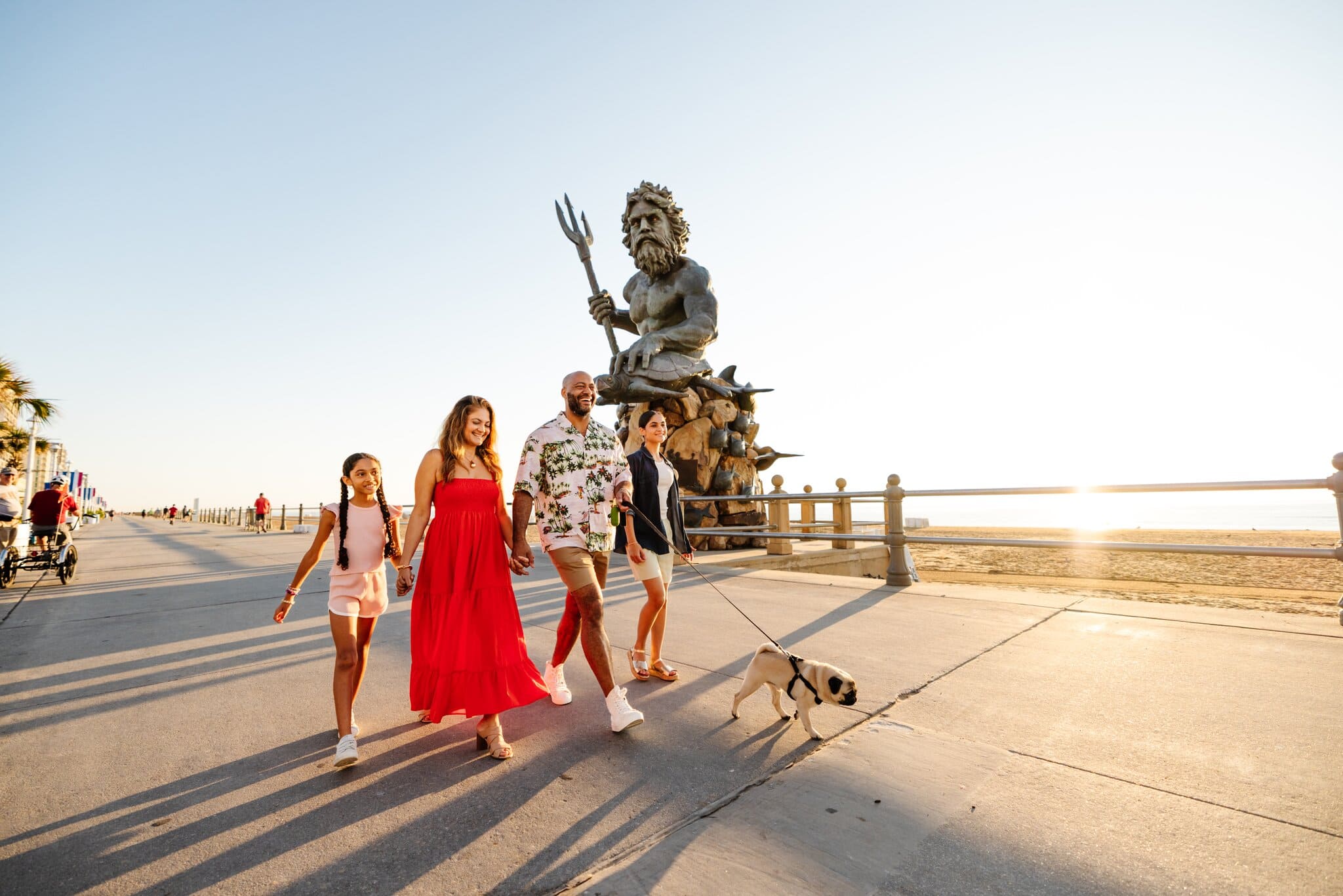 A family strolls along a beachfront promenade with a dog, passing by a large statue of a mythical figure at sunset.