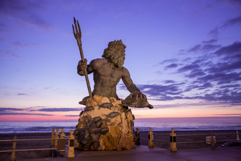 Statue of Poseidon holding a trident and a turtle, surrounded by sea creatures, against a twilight sky on a beach promenade.
