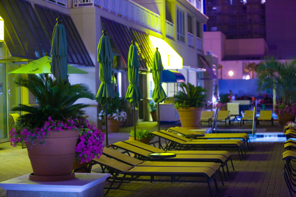 Poolside view at night with empty lounge chairs, green umbrellas, and large potted plants. Building lights illuminate the scene.