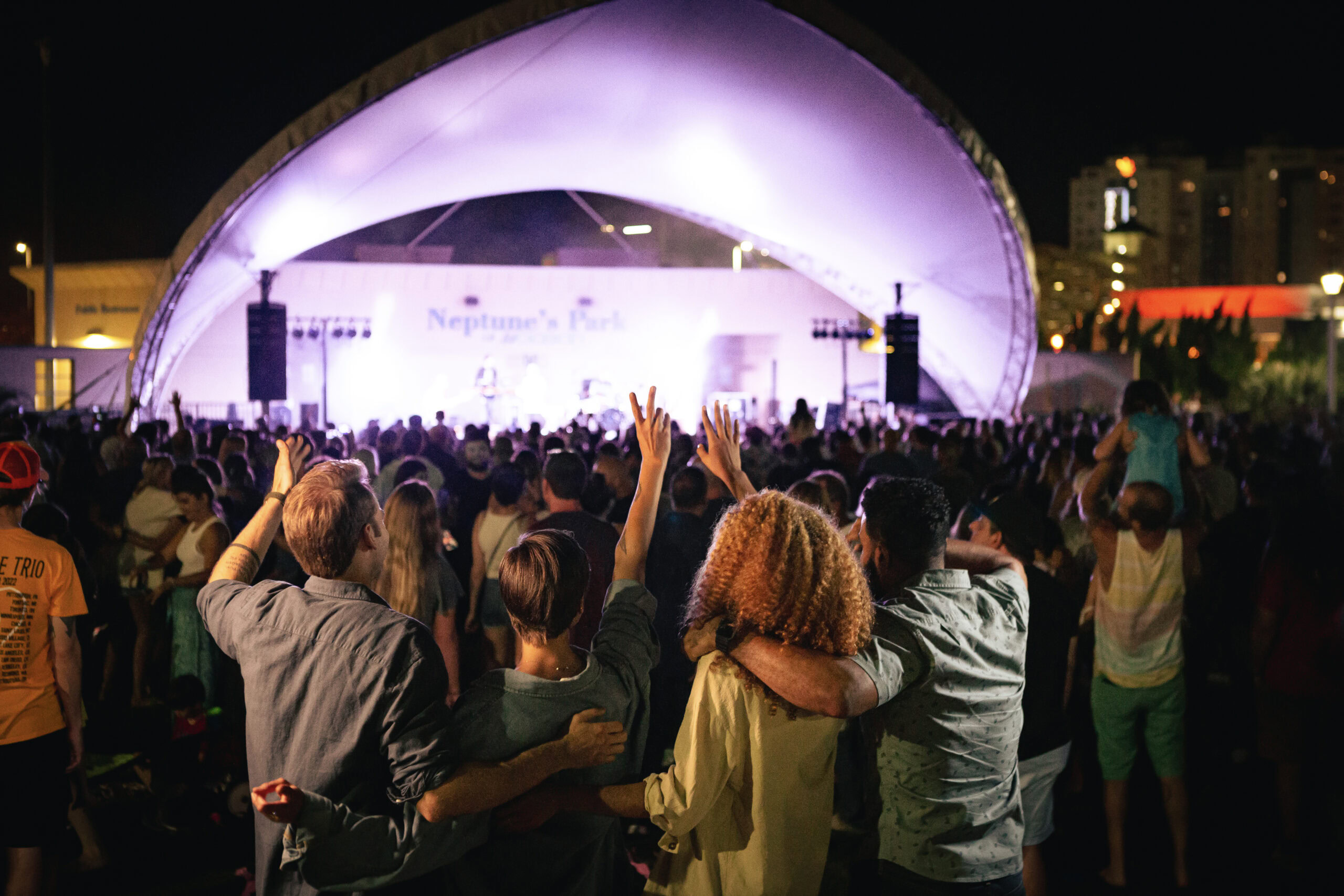 A crowd watches a live performance at an outdoor venue with a large, illuminated canopy. Some people are raising their hands, with buildings visible in the background at night.