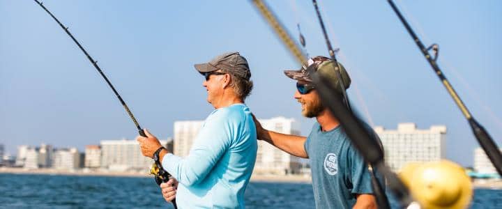 Two people fishing on a boat, smiling. City skyline in the background.