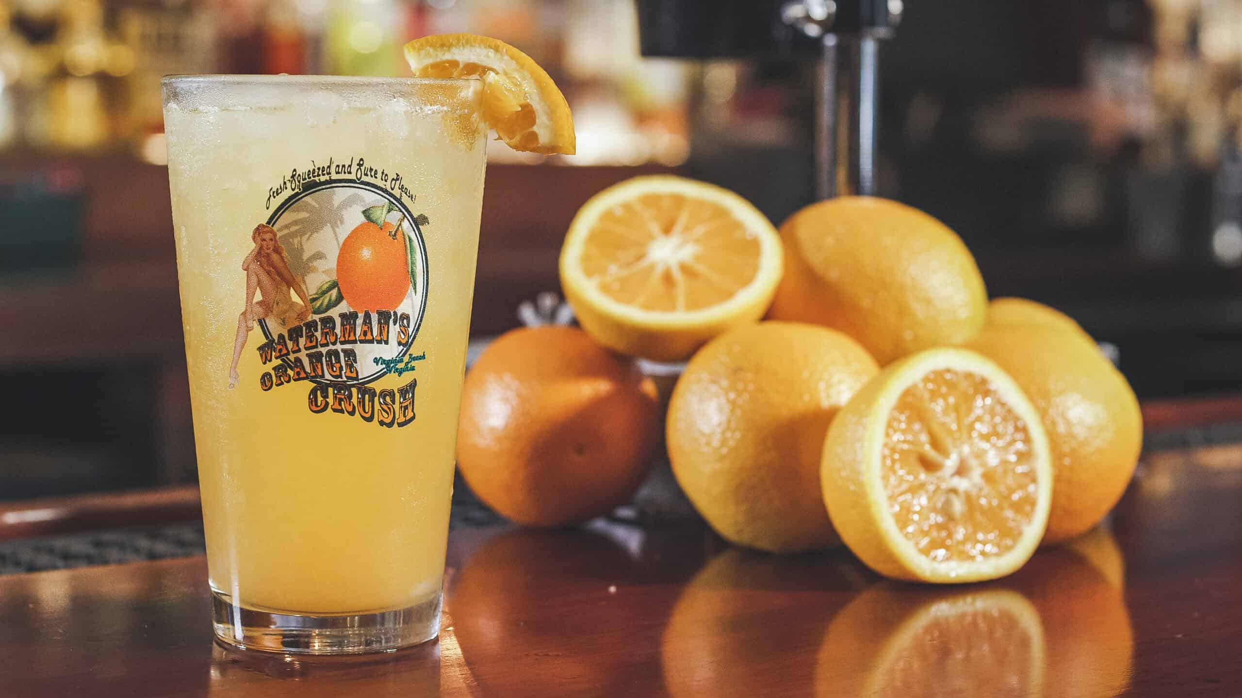 A frosty glass of orange juice with a lemon wedge on the rim sits on a bar next to a pile of whole and halved oranges.