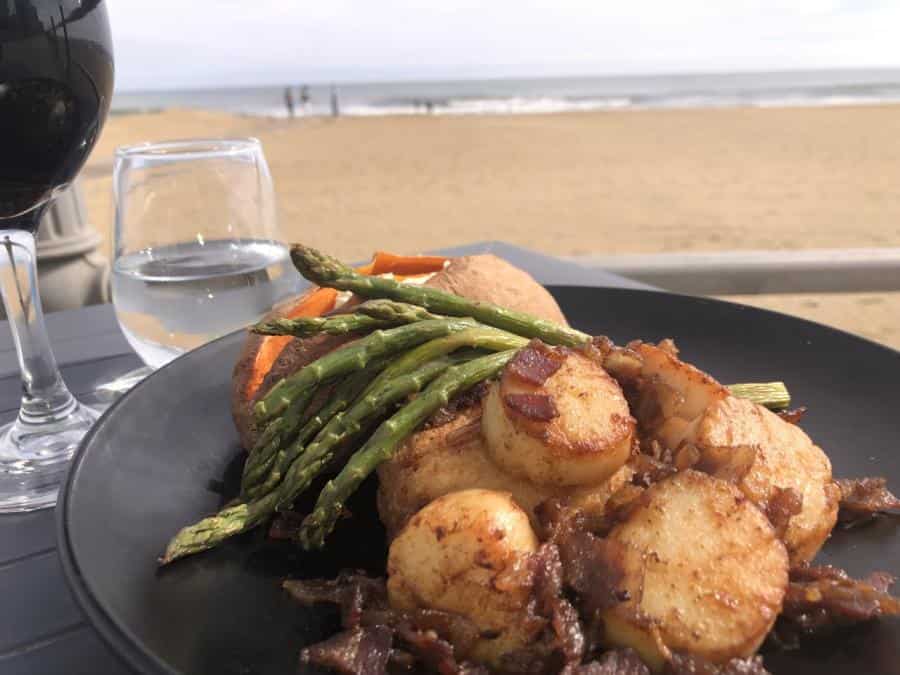 A plate of seared scallops with asparagus and onions, set on a beachside table. Accompanied by a glass of red wine and water, with the beach and ocean in the background.