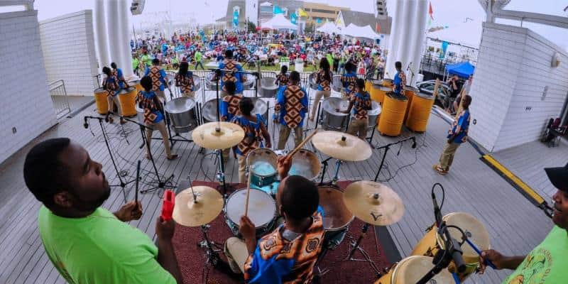 A band performs on stage with steel drums and a drum set. The audience is seated outdoors, some under tents. The musicians wear patterned outfits.