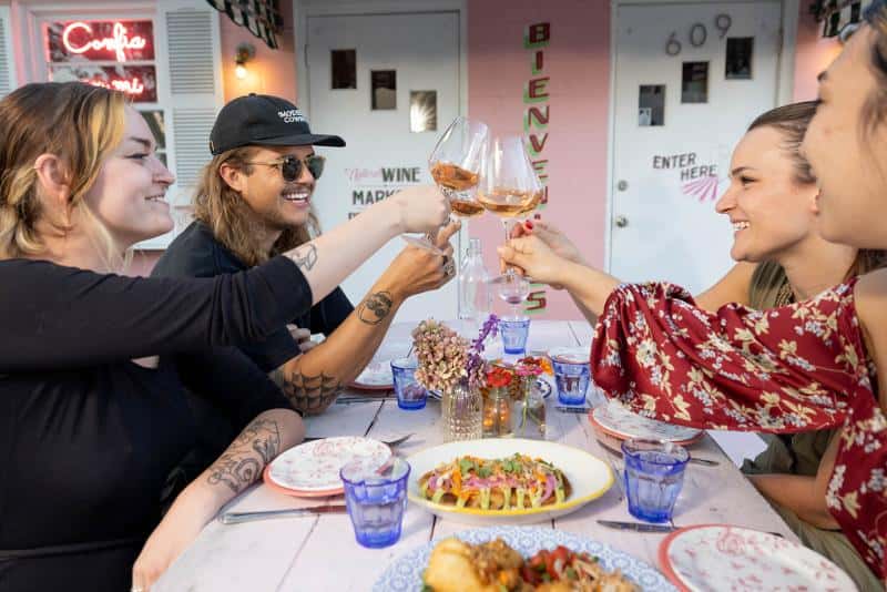 Four people sit at an outdoor table clinking wine glasses. Plates of food and blue glasses are on the table. A pink wall with "Bienvenidos" and house numbers is in the background.