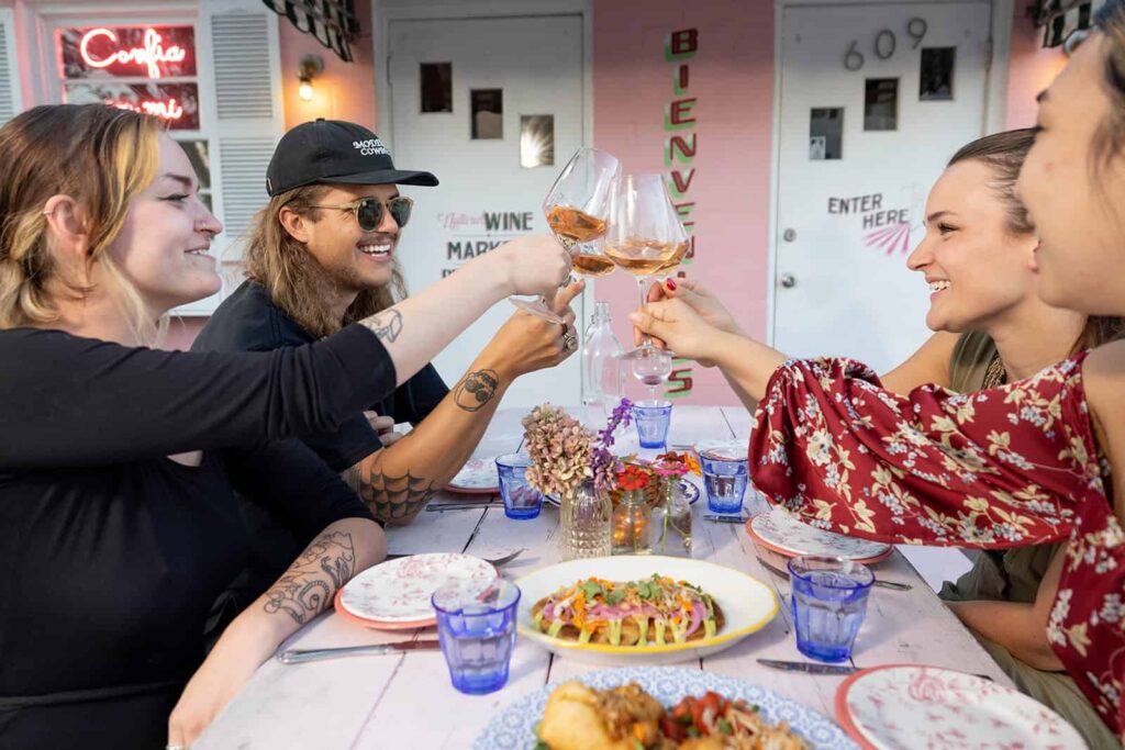 Four people sit at a table at the Pink Dinghy, toasting with wine glasses. Plates of food and blue glasses are on the table, with a pink walls in the background.