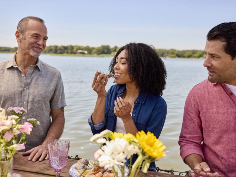 A diverse group of three friends enjoys a lakeside meal, sharing food and laughter among colorful flowers and the scenic water background.