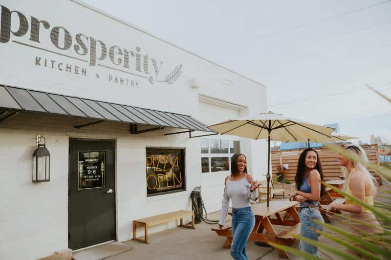 Three people stand and chat outside a restaurant named "Prosperity Kitchen + Pantry," with outdoor seating and a "Pizza Open" sign visible.
