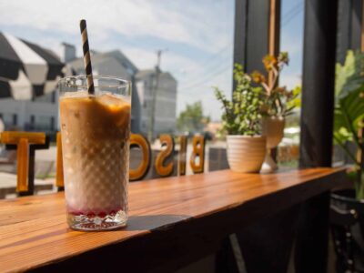 A glass of iced coffee with a black and white straw sits on a wooden table near a window, with plants and outdoor reflections in the background.