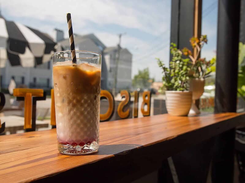 A glass of iced coffee with a black and white straw sits on a wooden table near a window at Three Ships Coffee, with plants and outdoor reflections in the background.