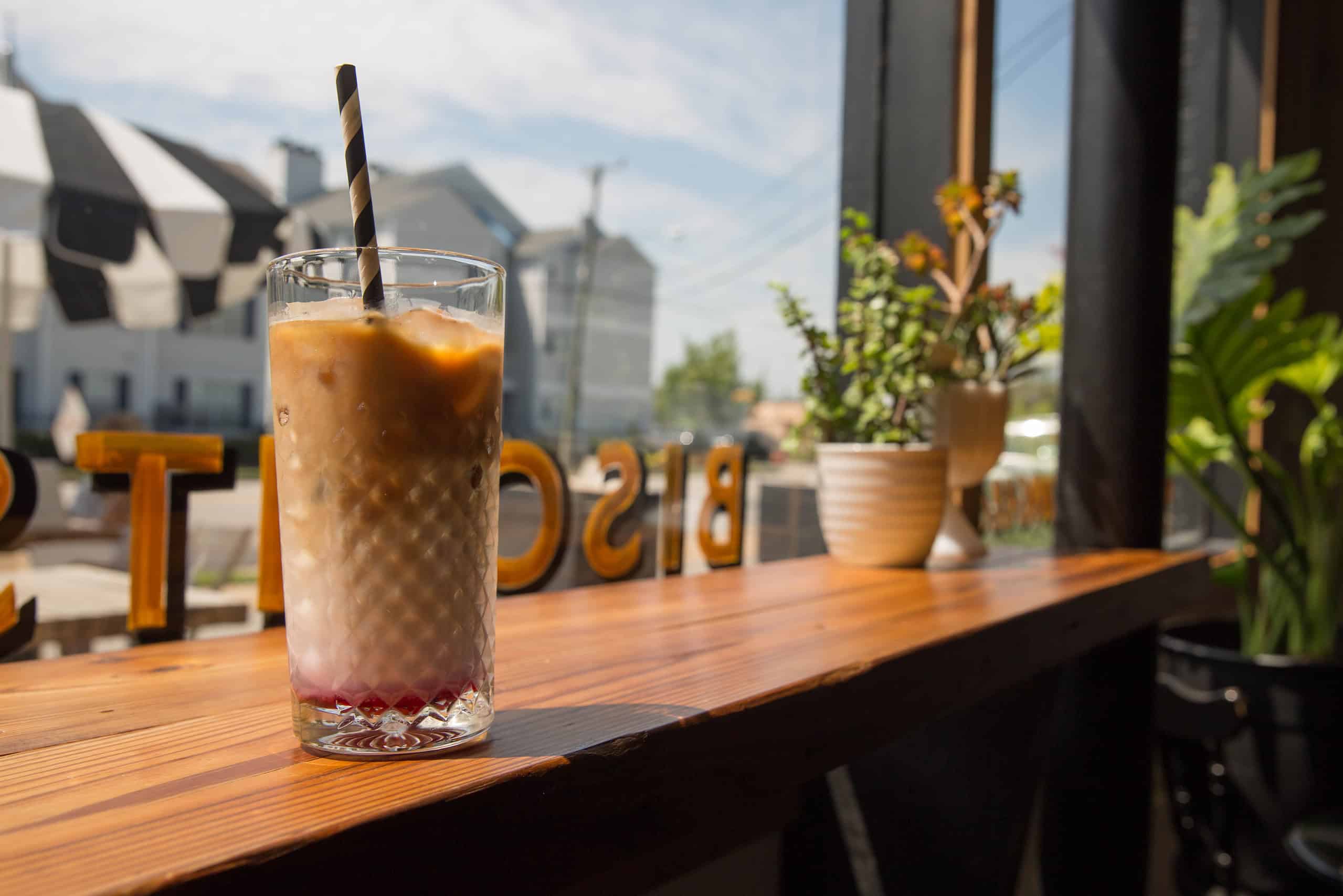 A glass of iced coffee with a black and white straw sits on a wooden table near a window at Three Ships Coffee, with plants and outdoor reflections in the background.
