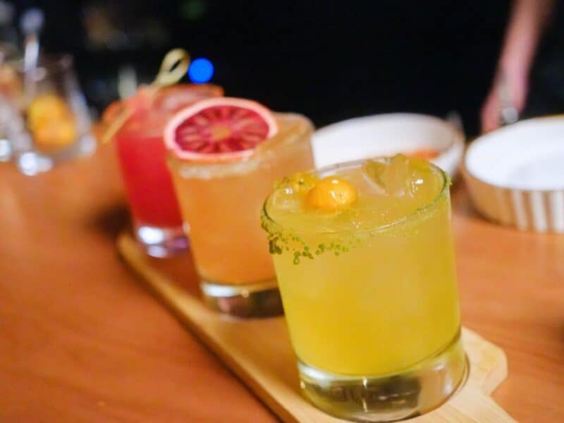 A wooden tray displays three colorful cocktails, including a vibrant yellow drink with a garnish, a blood orange cocktail, and a pink drink, set against a blurred background.