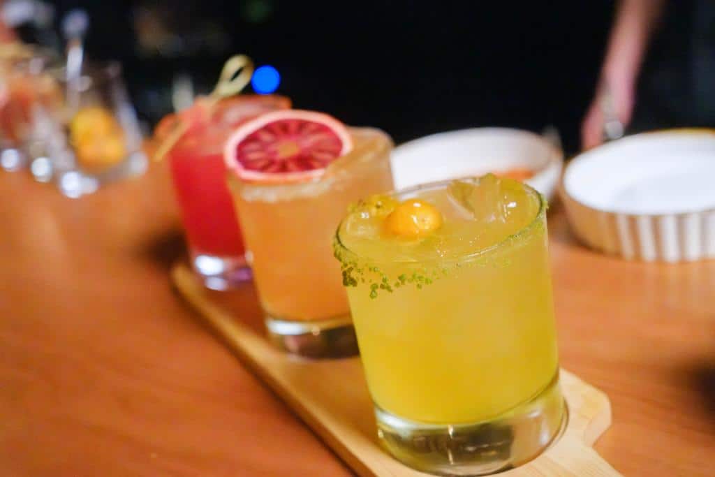 A wooden tray displays three colorful cocktails, including a vibrant yellow drink with a garnish, a blood orange cocktail, and a pink drink, set against a blurred background.