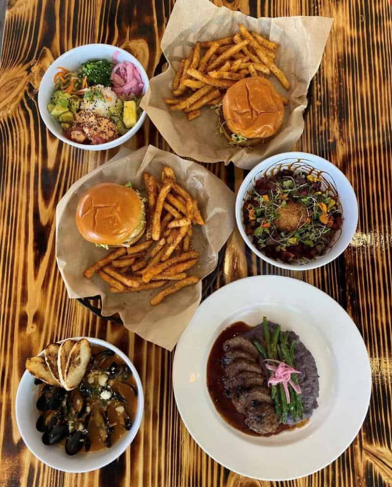 Top-down view of a wooden table with two burgers and fries, a poke bowl, a mussel dish, a salad, and a steak plate with asparagus and mashed potatoes.