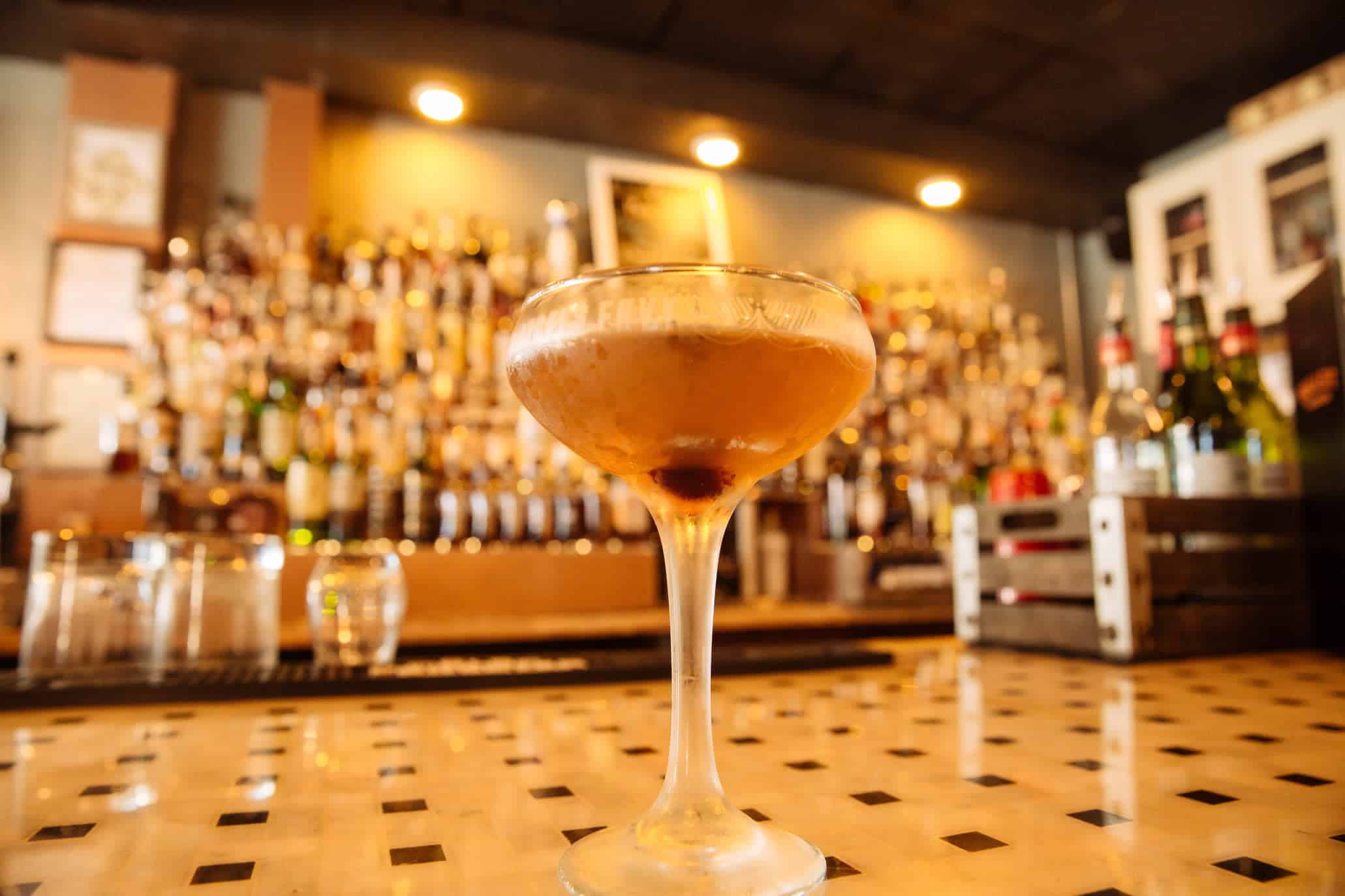 A cocktail in a coupe glass sits on a bar counter, with a blurred background of various bottles and dim lighting.