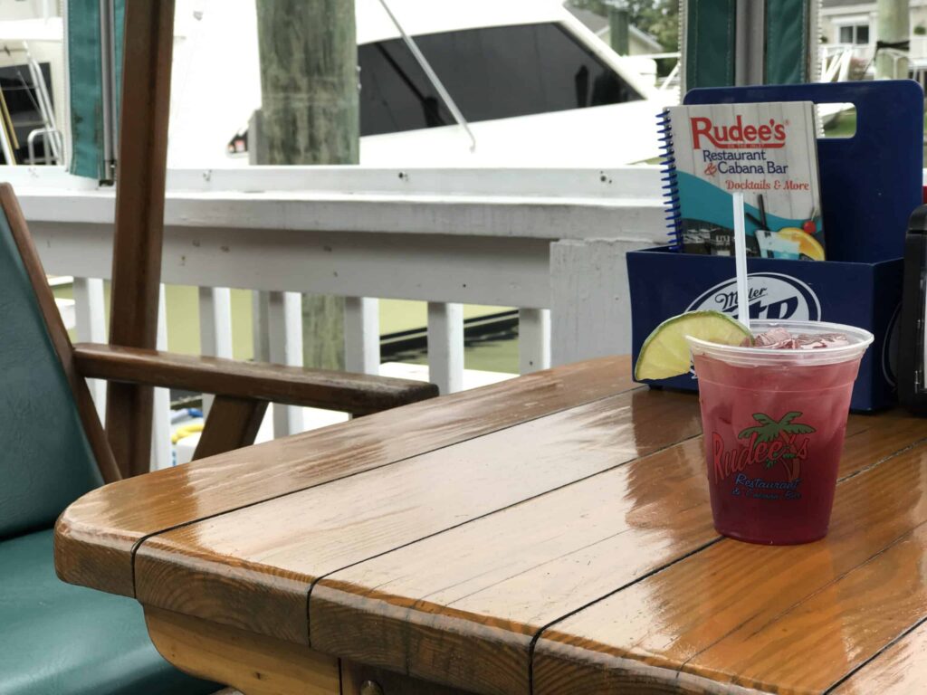 A red drink with a lime wedge sits on a wooden table at a marina restaurant. A menu and docked boat are in the background.