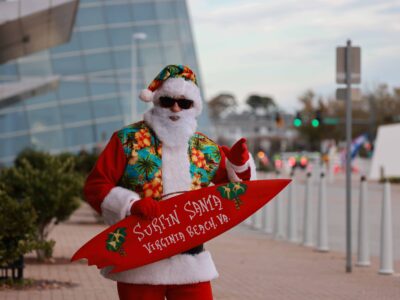 Person dressed as Santa in tropical attire, holding a red surfboard with 