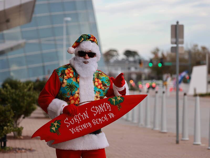 Person dressed as Santa in tropical attire, holding a red surfboard with "Surfin Santa Virginia Beach, VA" written on it, standing on a sidewalk near a modern building.