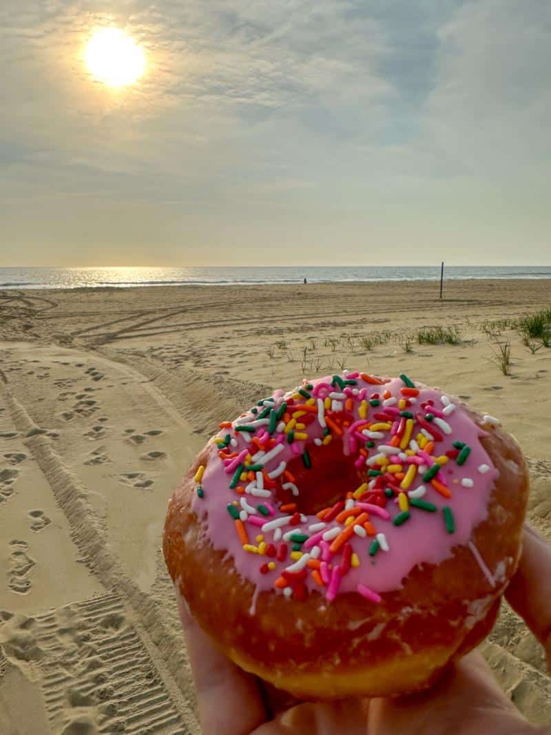 A donut with pink icing and colorful sprinkles is held up in front of a sandy beach and ocean, with the sun low in the sky.