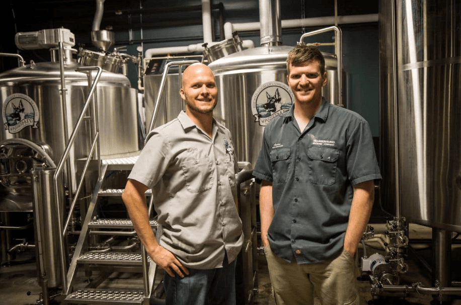 Two men stand in front of large stainless steel brewing tanks inside a brewery, both wearing short-sleeve work shirts and smiling at the camera.
