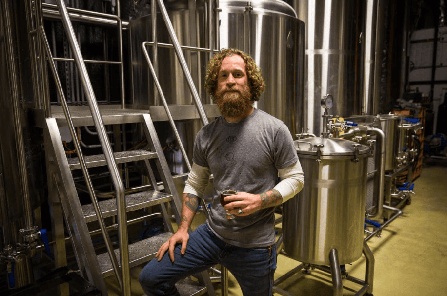 A man with a beard stands next to stainless steel brewing equipment, holding a glass, in an industrial brewery setting.