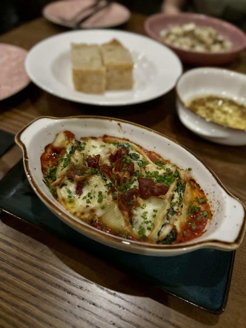 A dish of stuffed pasta shells with spinach, cheese, and garnished with crispy prosciutto, served in a white oval dish on a wooden table, with bread and olive oil in the background.