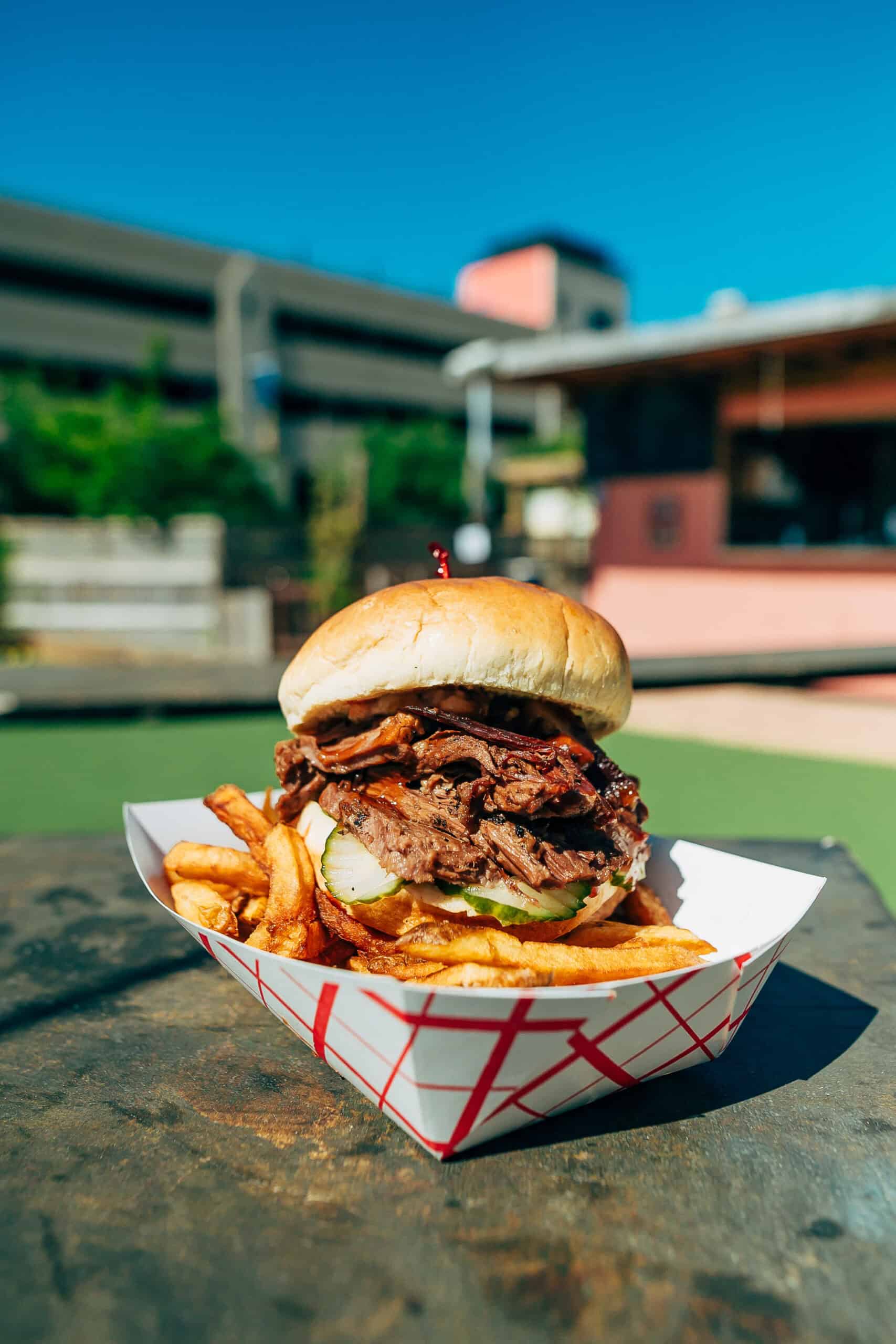 A large beef sandwich with caramelized onions, lettuce, and pickle in a white paper tray, accompanied by fries.