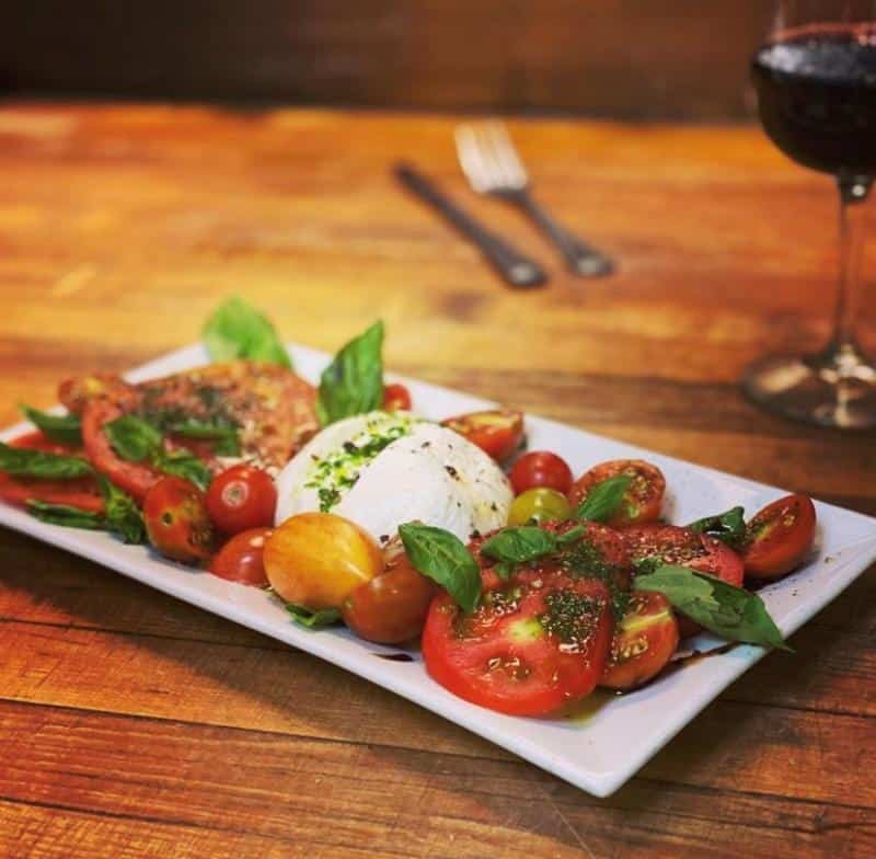 A plate with fresh mozzarella, sliced tomatoes, cherry tomatoes, basil, and seasoning, on a wooden table with a glass of red wine in the background.
