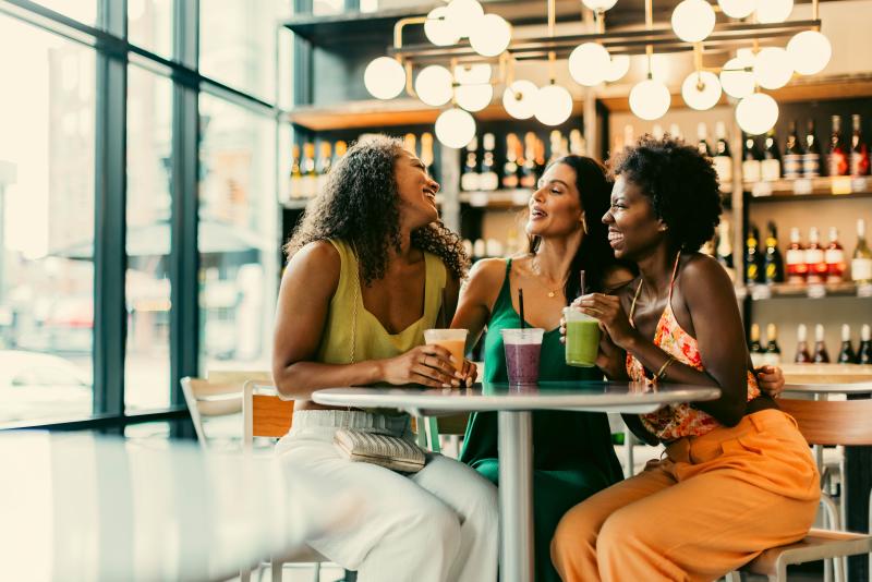 Three women sit laughing at a café, each holding a colorful drink. Bottles and bright lights are in the background.