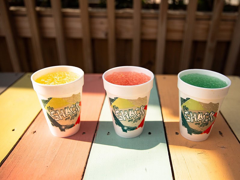Three Styrofoam cups with colorful frozen drinks—yellow, red, and blue—are placed on a pastel wooden table outdoors.