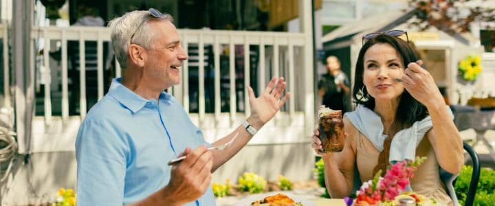 A man and woman sit at an outdoor cafe table. The man gestures with his hands, while the woman holds a fork and a dessert. They appear to be engaged in conversation.