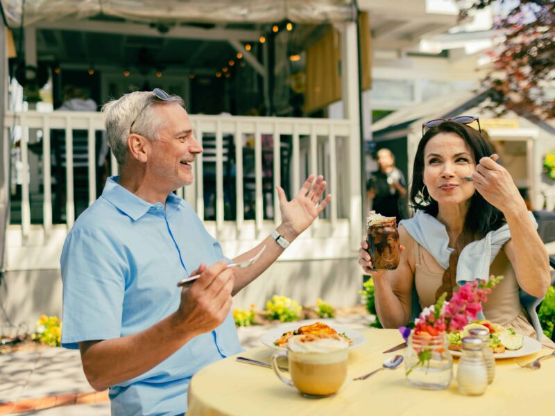 A man and woman sit at an outdoor cafe table. The man gestures while smiling, and the woman eats dessert with a spoon. There is food and drinks on the table.