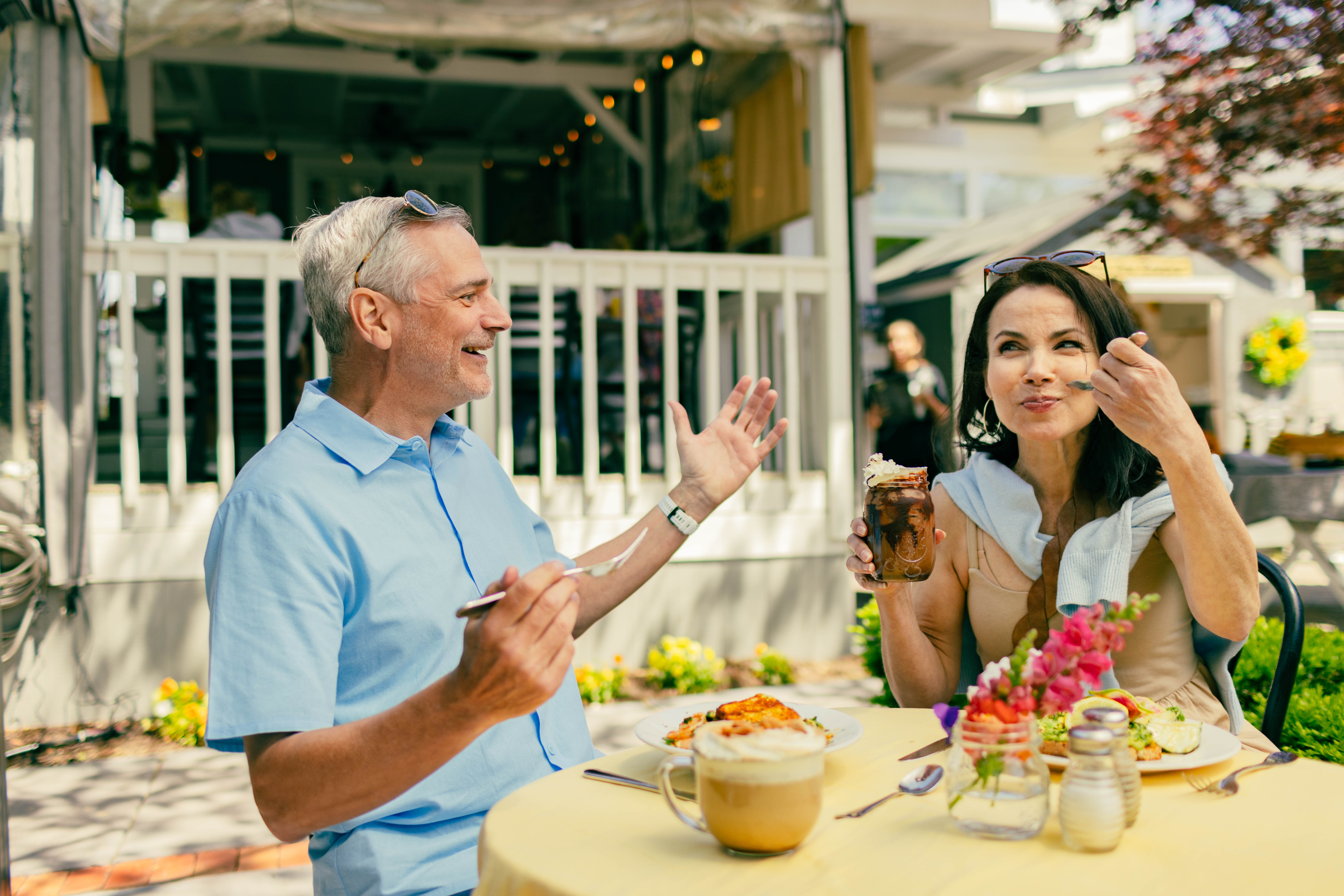 Man and woman enjoying a meal at an outdoor cafe table. Woman is holding a dessert, while the man gestures with a fork. Bright, sunny ambiance.