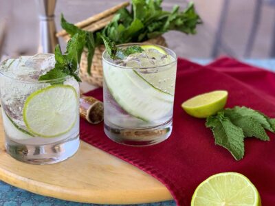 Two glasses of clear cocktails with lime slices and mint, on a wooden board. A basket of mint, a halved lime, and mint leaves are on a red cloth nearby.