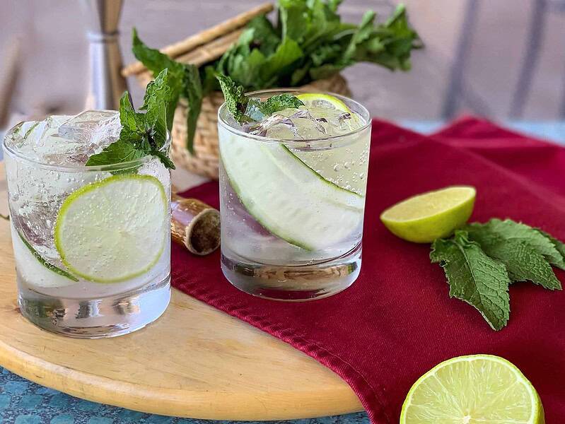 Two glasses of clear cocktails with lime slices and mint, on a wooden board. A basket of mint, a halved lime, and mint leaves are on a red cloth nearby.