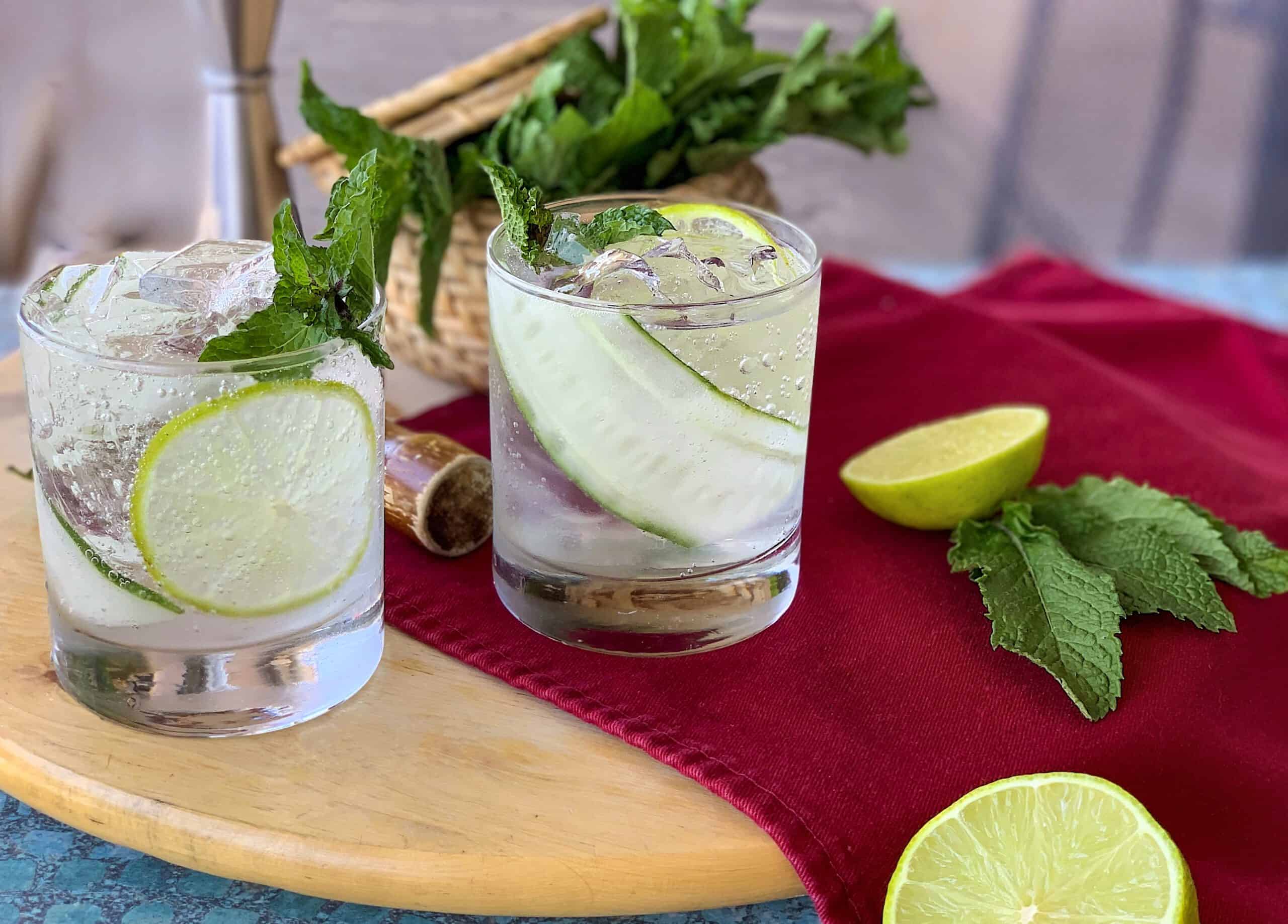 Two glasses of clear cocktails with lime slices and mint, on a wooden board. A basket of mint, a halved lime, and mint leaves are on a red cloth nearby.