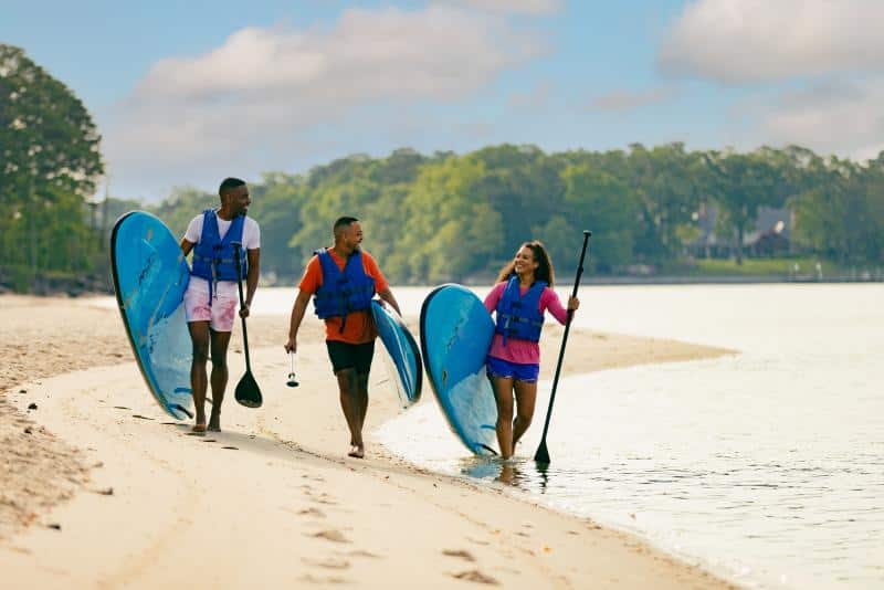Three people in life vests walk along a sandy beach carrying blue stand-up paddleboards near a calm body of water. Trees are visible in the background.