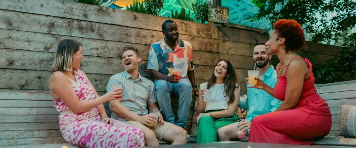 Seven people are seated on an outdoor bench, holding drinks and laughing, surrounded by greenery and wooden paneling.