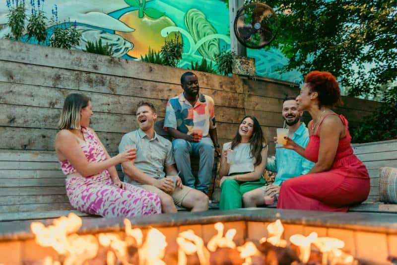 A group of six people sit and stand around a fire pit, laughing and holding drinks, with a mural and plants in the background.
