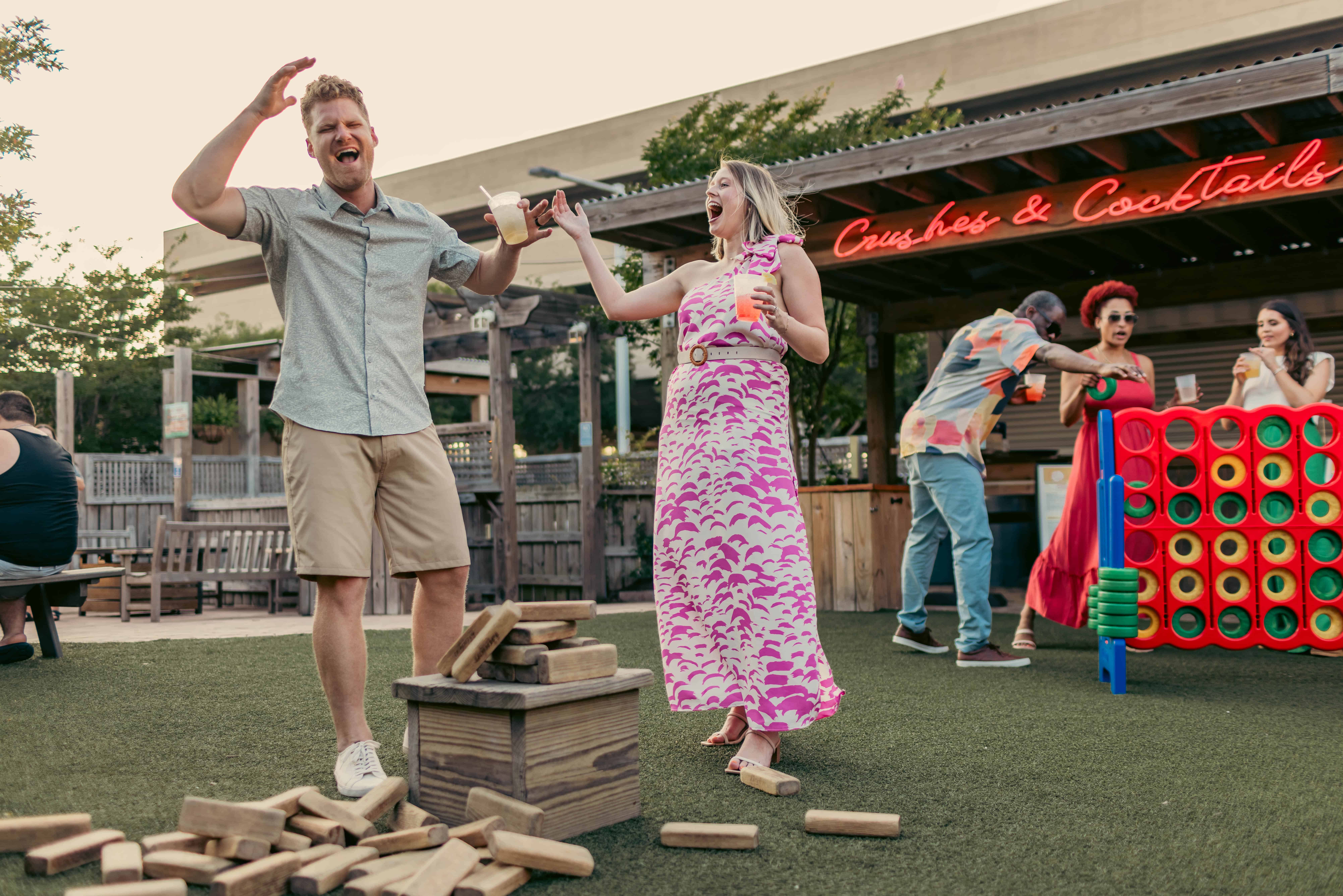 Man and woman laugh while playing a stacking block game outdoors. People in the background enjoy drinks near a large Connect Four game.