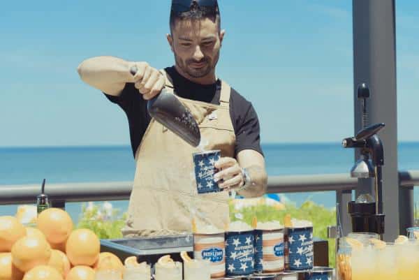 Man in an apron pours a drink into a cup at an outdoor bar with oranges and drink containers. Ocean visible in the background.