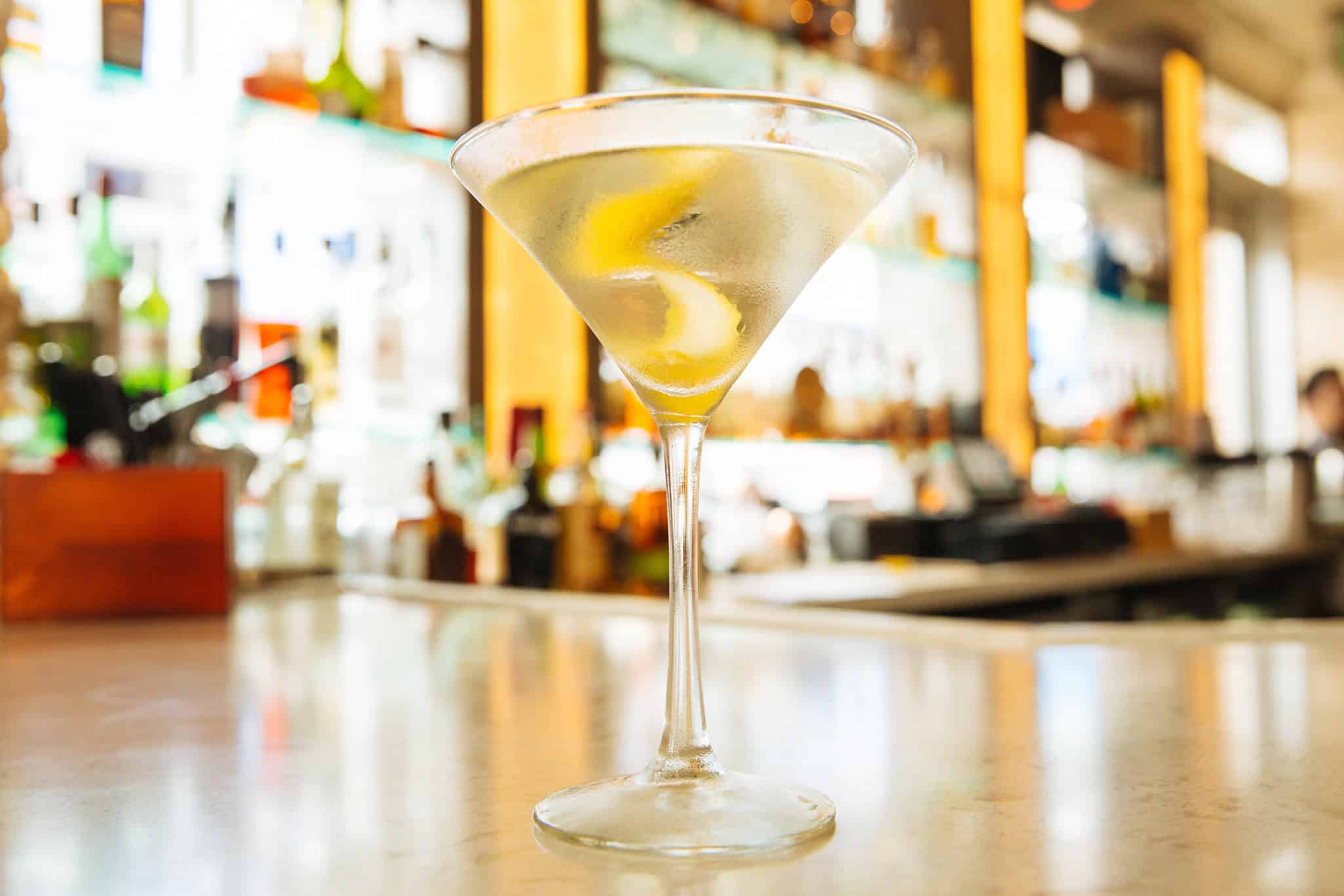 A martini glass with a clear drink and lemon twist on a bar counter with blurred bottles in the background.