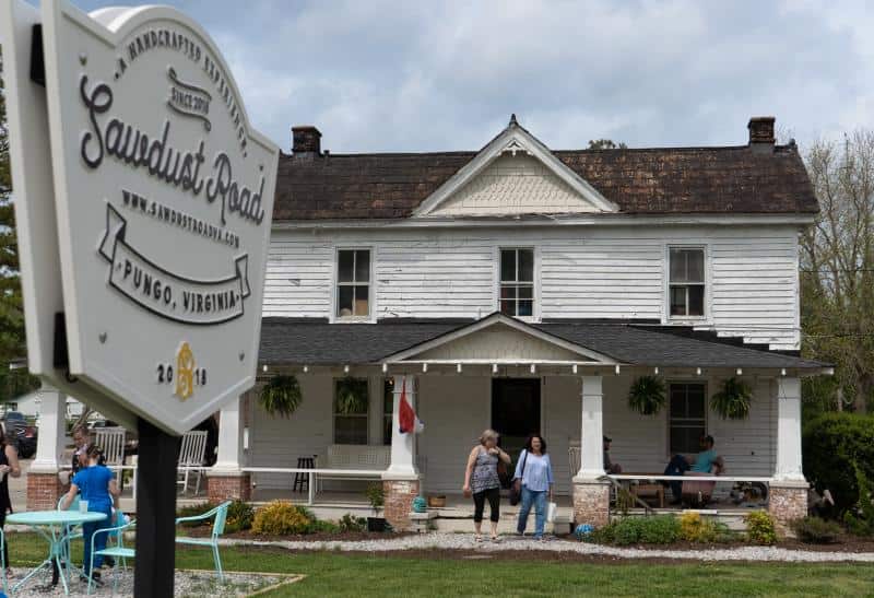 A weathered white two-story house with a sign in the foreground that reads "Sawdust Road, Pungo, Virginia." Several people are on the porch and near the front door.
