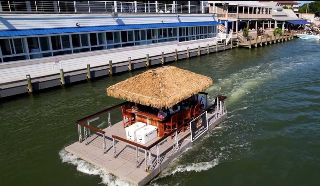 A tiki bar boat with a thatched roof floats on a waterway, heading away from a dock next to a waterside building.