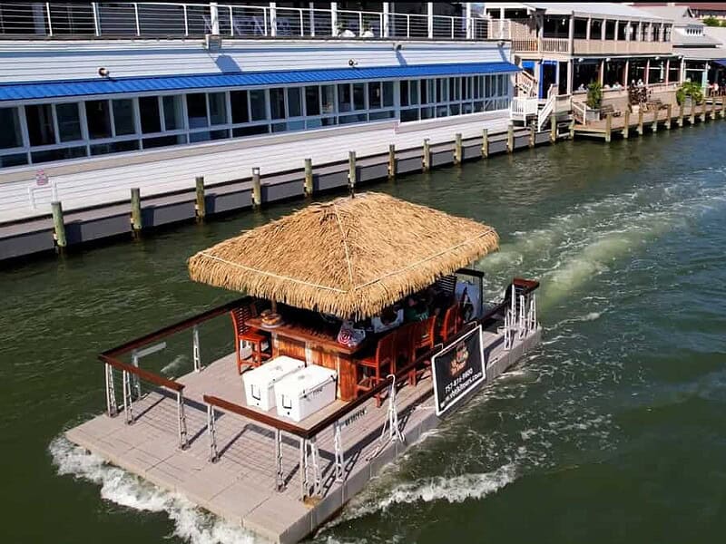 A tiki bar boat with a thatched roof floats on a waterway, heading away from a dock next to a waterside building.