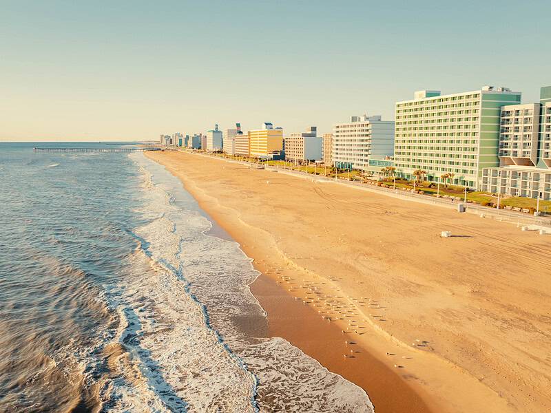 Coastal landscape featuring sandy beach, gentle ocean waves, and modern beachfront buildings under a clear sky.