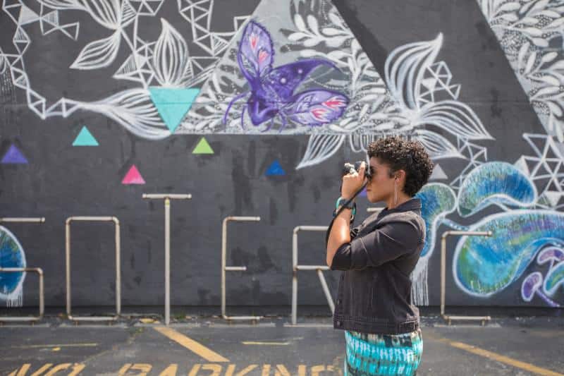 Person taking a photo in front of a vibrant mural with geometric shapes and a butterfly.