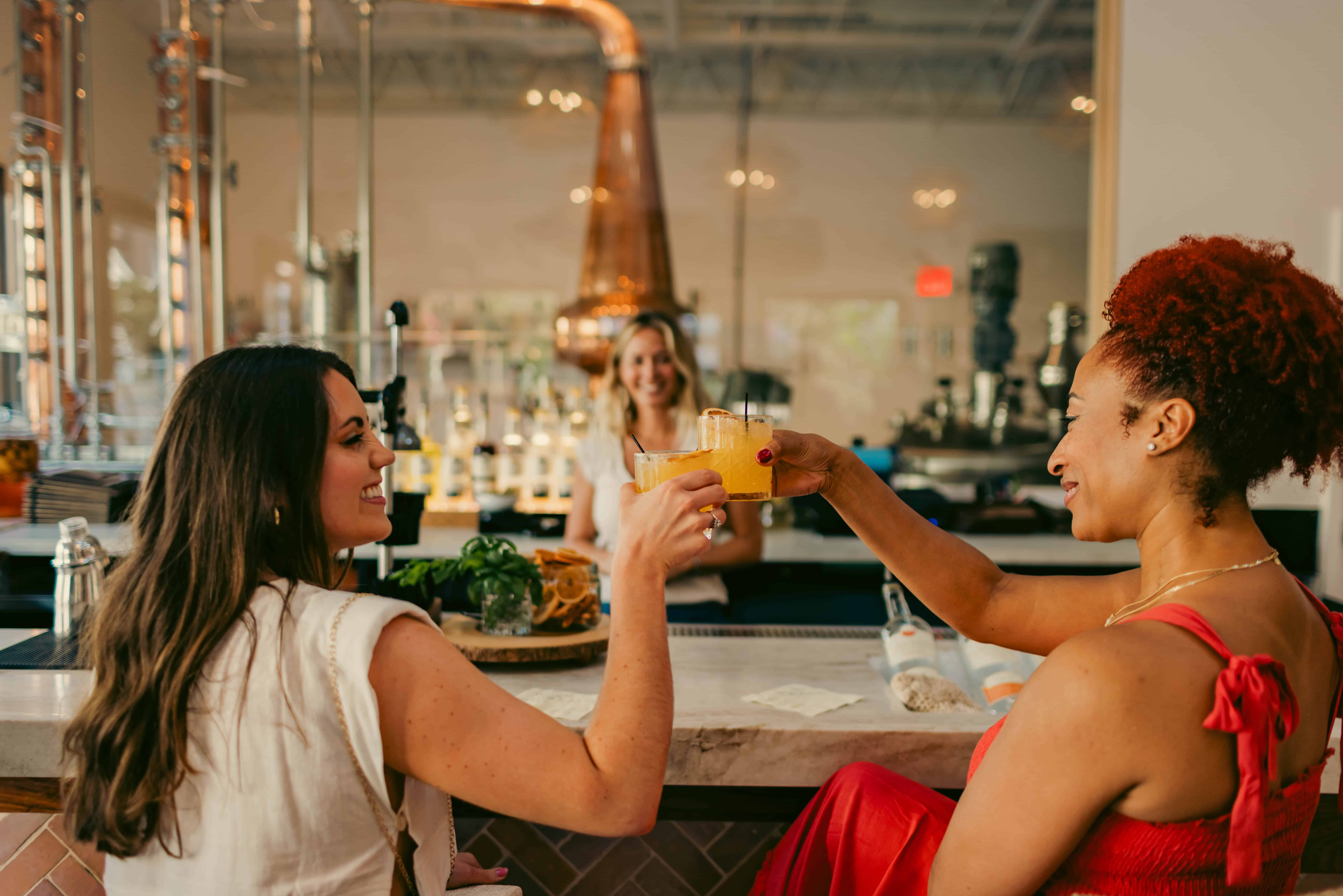 Two women clink glasses at a bar, sitting on stools. A bartender smiles in the background.