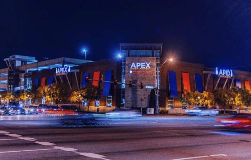 Night view of a modern entertainment complex with "APEX" signs, illuminated against a dark sky. Cars are visible on the road in front.