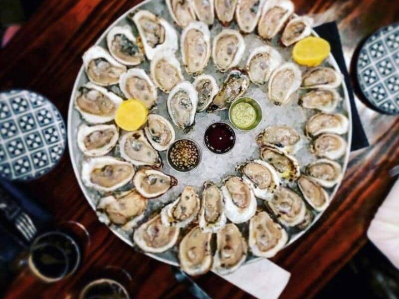 A large platter of oysters on ice is surrounded by lemon wedges, dipping sauces, and seasoning, set on a wooden table with patterned plates visible at the edges.